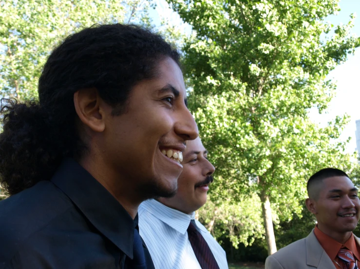 two men smile while standing outside next to trees