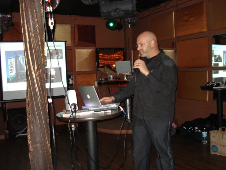 a bald man in glasses stands by a microphone near a laptop and speaks