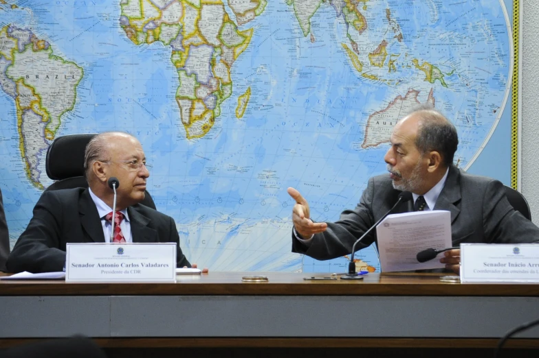 two men sitting at a desk talking and gesturing