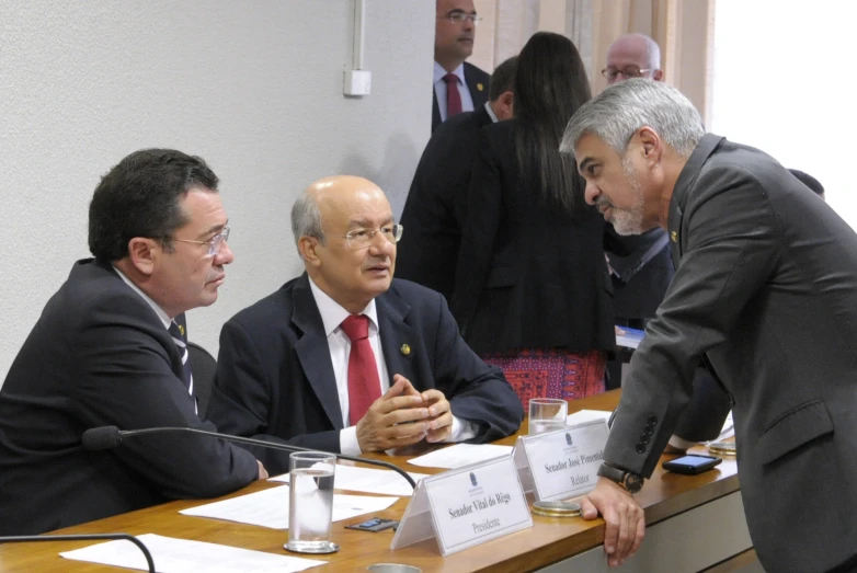 three men sitting in front of microphones at a meeting