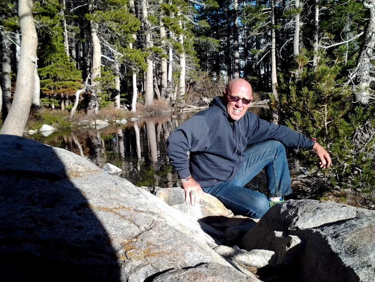 a man in sunglasses crouching on rocks with trees and mountains in the background