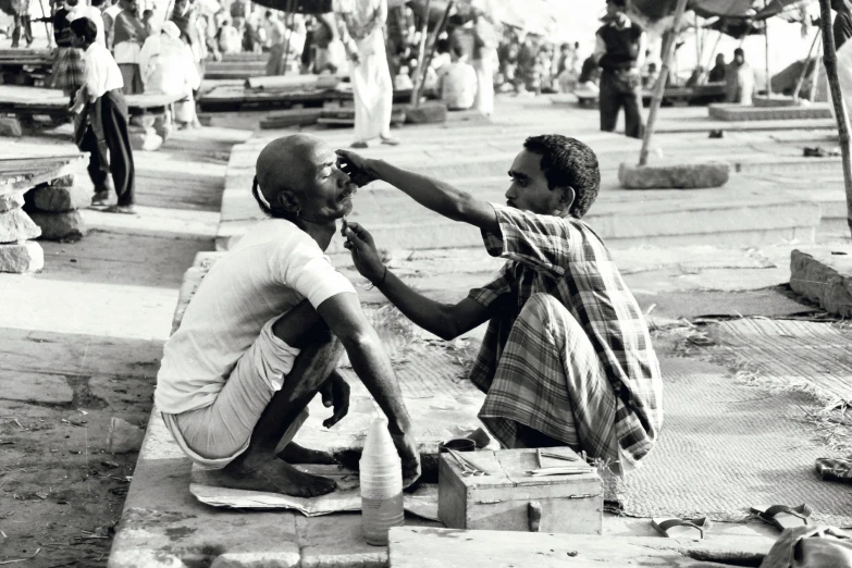 two men are sitting on the ground, one is combing the other's hair