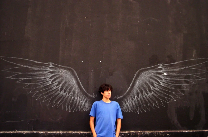 a boy stands next to a chalk board wall with wings