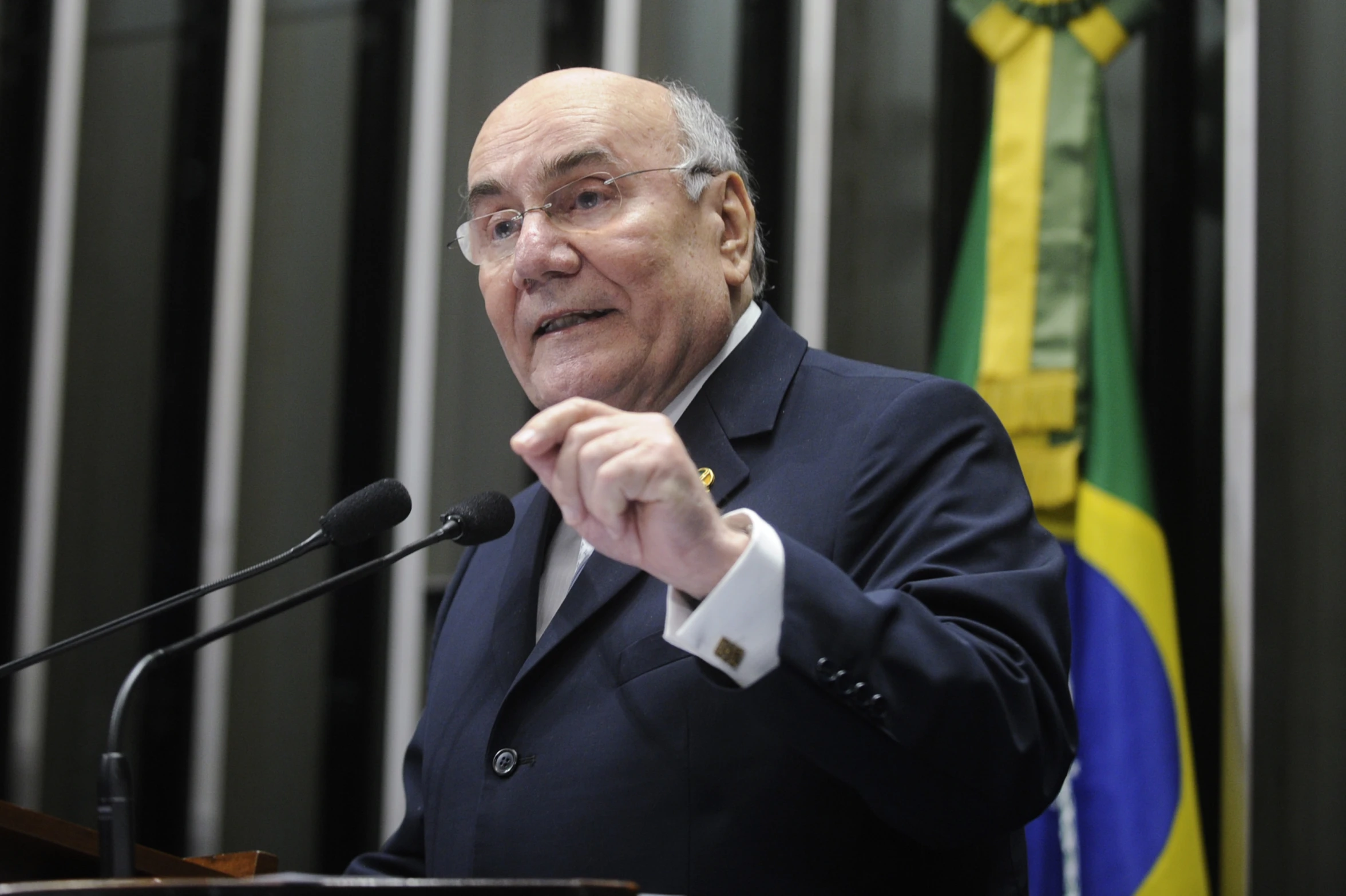 a man wearing glasses and suit speaking at a podium