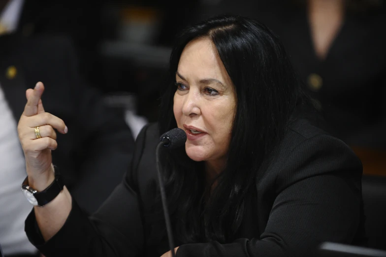 a woman in a black jacket holds her hand up to her right as she sits in front of a microphone