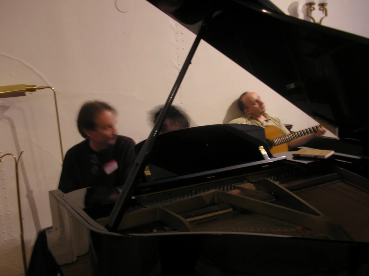 two men sit at a piano, one with a guitar in his lap