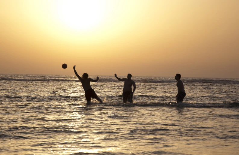 three men playing beach volleyball in the water at sunset