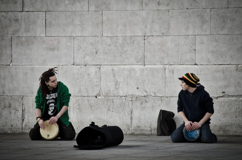 two men sit on the ground near wall with their instruments