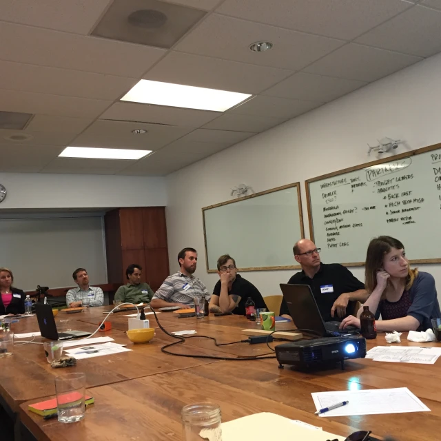 a group of people sitting at a long table with laptops on it