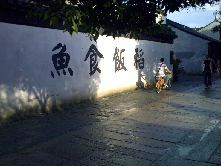 the person on the bike is walking beside a wall with chinese writing