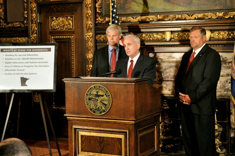 three men wearing ties stand at podiums