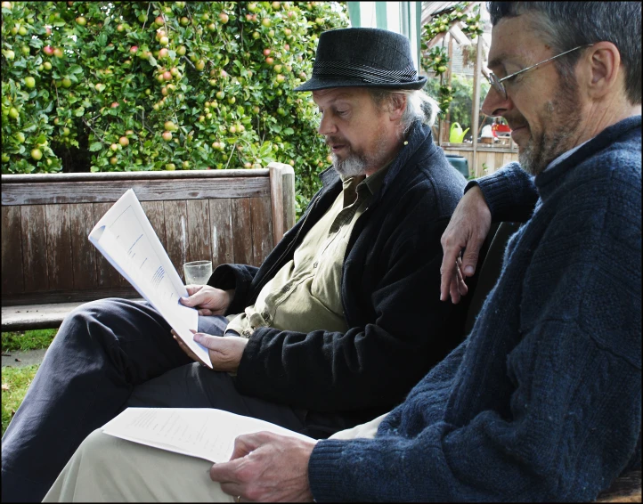 two older men are looking at books on a park bench