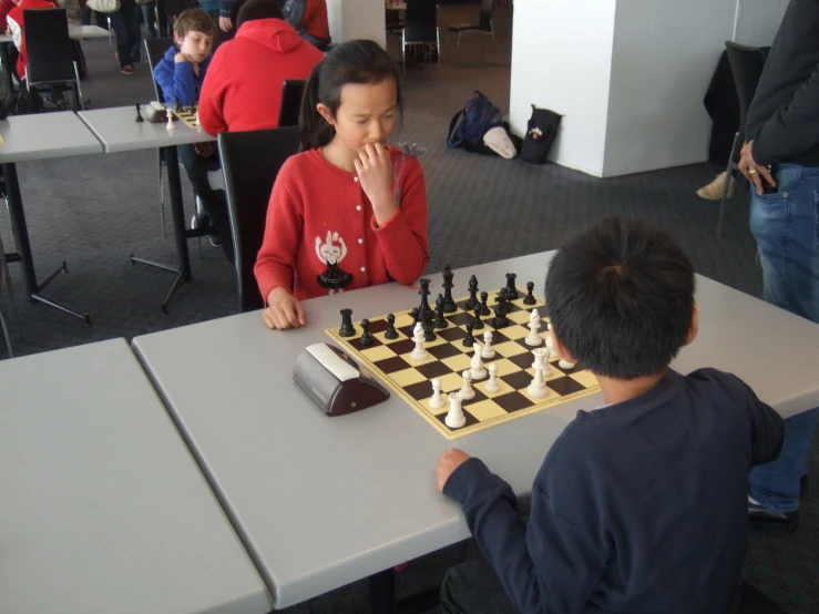 an asian boy sitting at a table next to his sister, who is playing chess