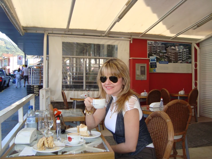 a woman in sunglasses sitting down at an outdoor restaurant drinking tea