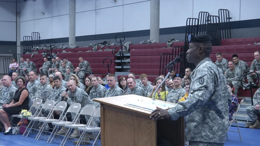 an army man in camouflage stands in front of a podium while other soldiers sit and talk