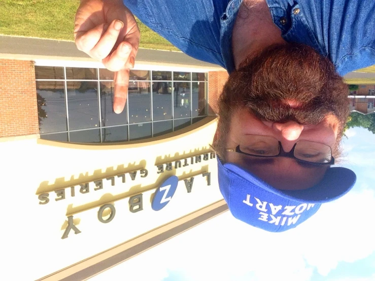 a bearded man with glasses and a blue cap is outside a clothing store and pointing the middle finger