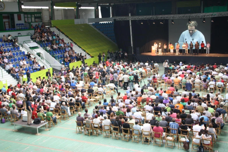 an auditorium full of people, people sitting in chairs with the speaker projected