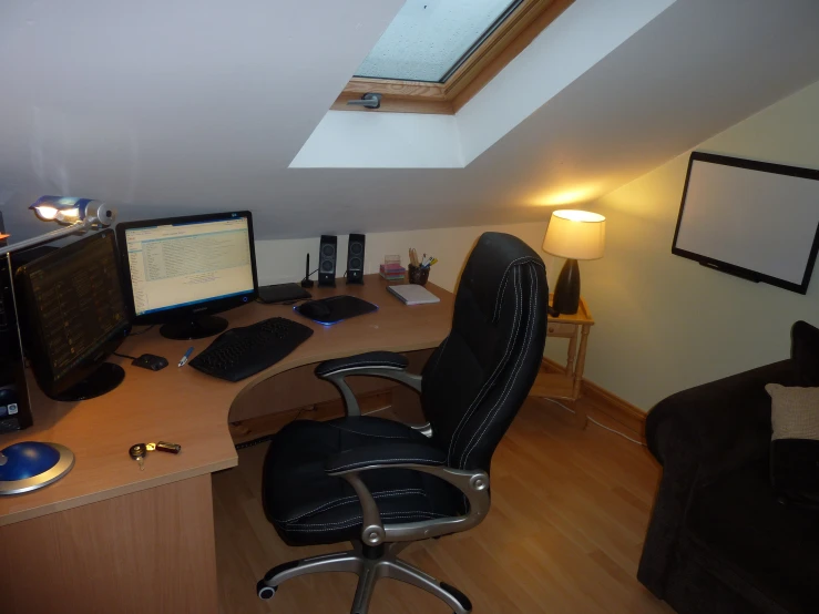 a office room has a slanted ceiling, with a desk, chair, computer, and monitor