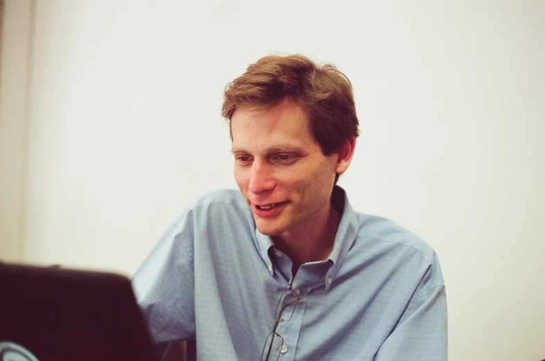 a man with long brown hair in a blue shirt smiles while sitting in front of a laptop computer