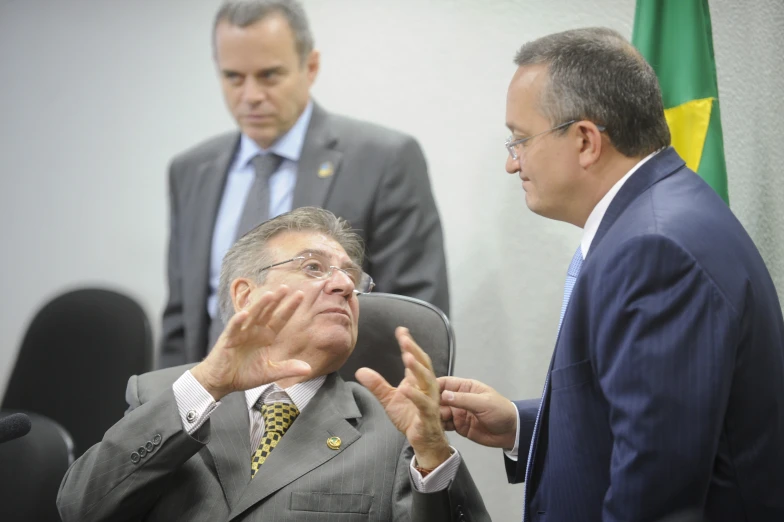 two men in suits standing and sitting near each other