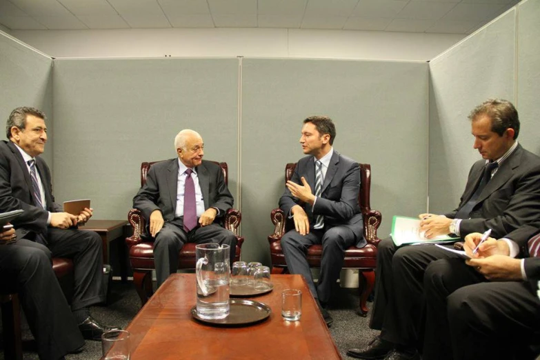 men sitting and talking in chairs with water, papers, and notebooks on a table