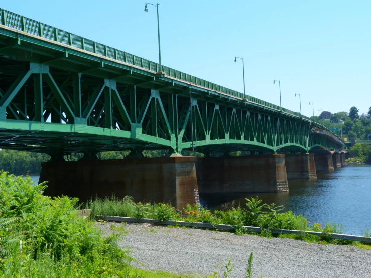 a green metal bridge over water with grass and trees