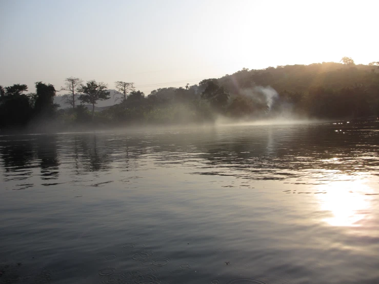 trees line the banks of a lake as the sun is seen shining over it