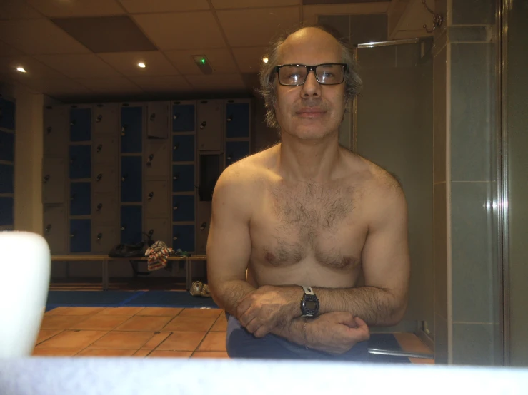 a bald man with glasses sits in a bathroom, wearing an arm watch