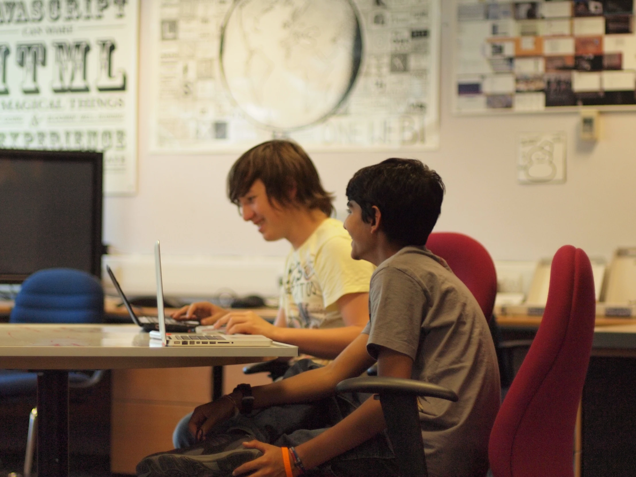 two boys sitting at a table working on their laptops