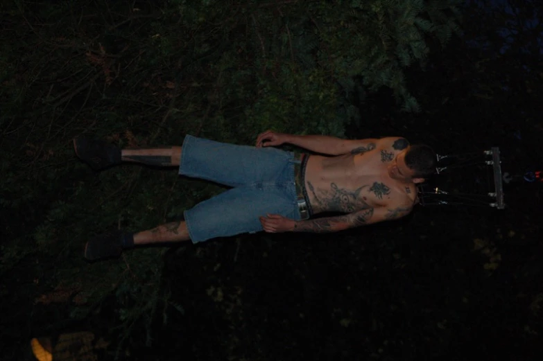 a man with tattoos, jeans shorts and an electric chair suspended from a string in the dark