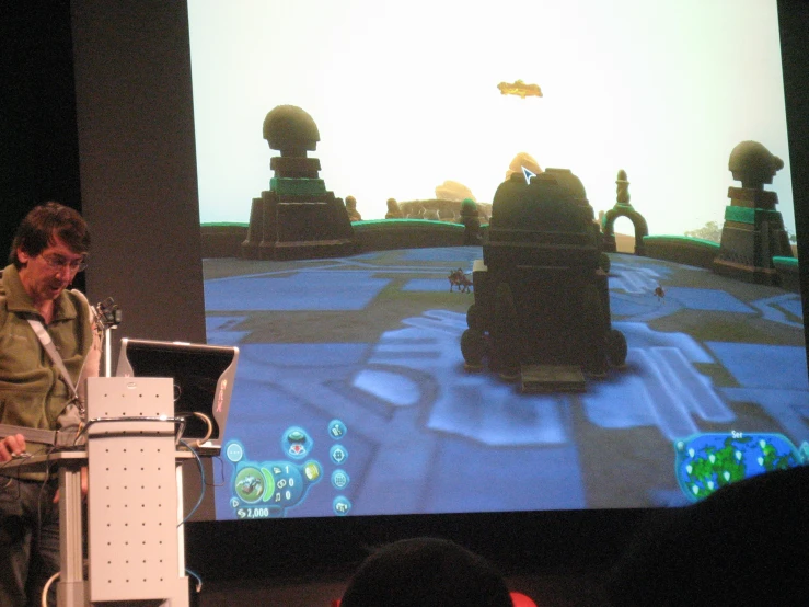 man standing at podium with projection screen showing game in background