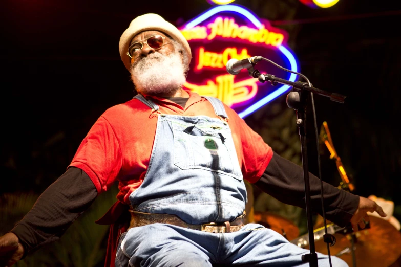 an older man sitting on a stool while singing into a microphone