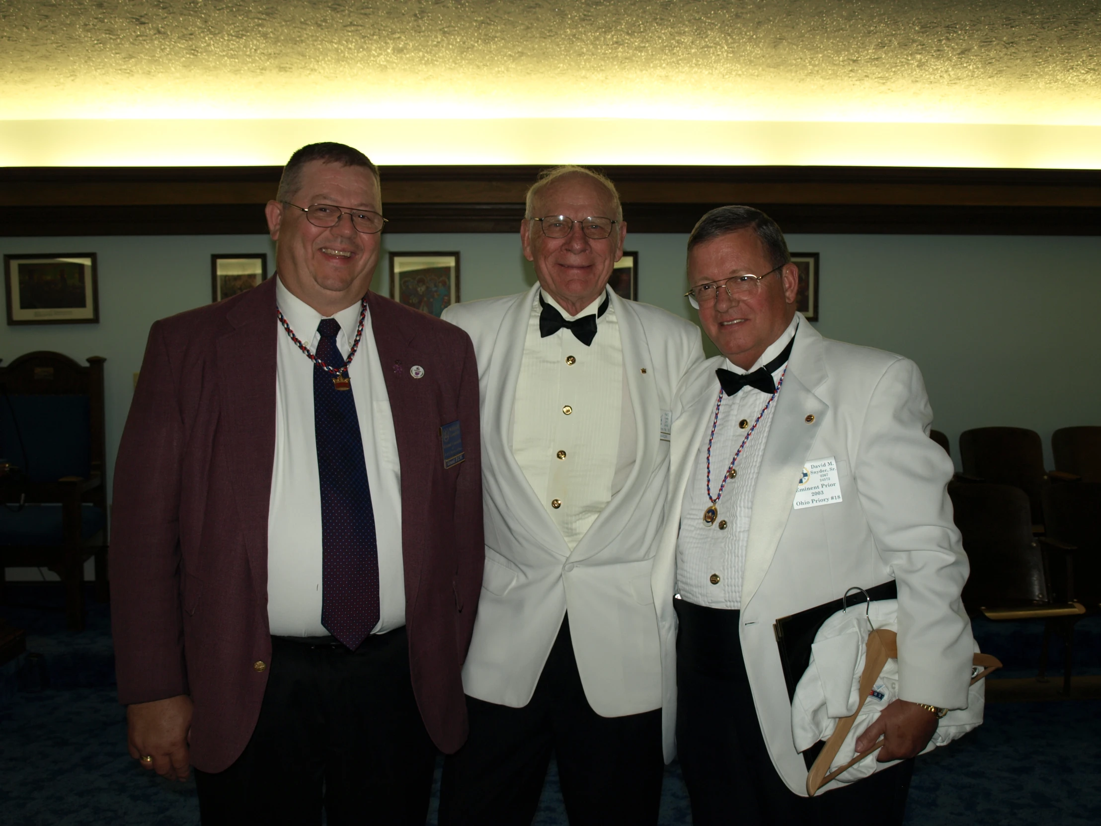 two older men wearing tuxedo suits and holding pocket watches stand beside a younger man in glasses