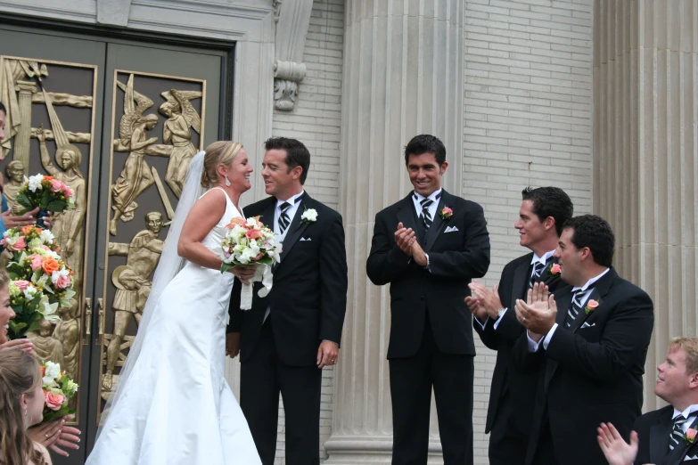 a bride and groom in wedding suits are greeting the bride in front of a statue