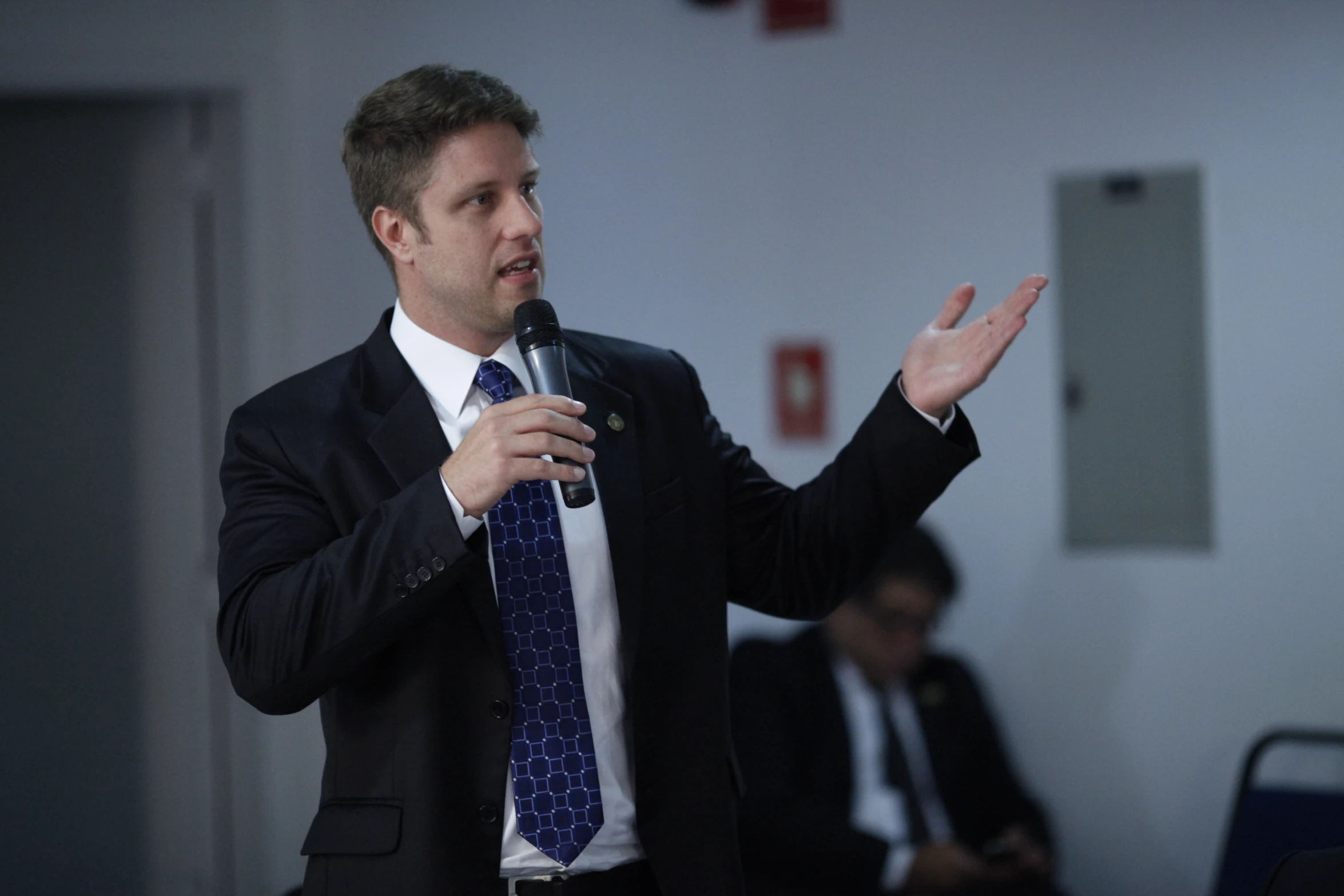 man speaking with microphone and suit, on stage with three people sitting