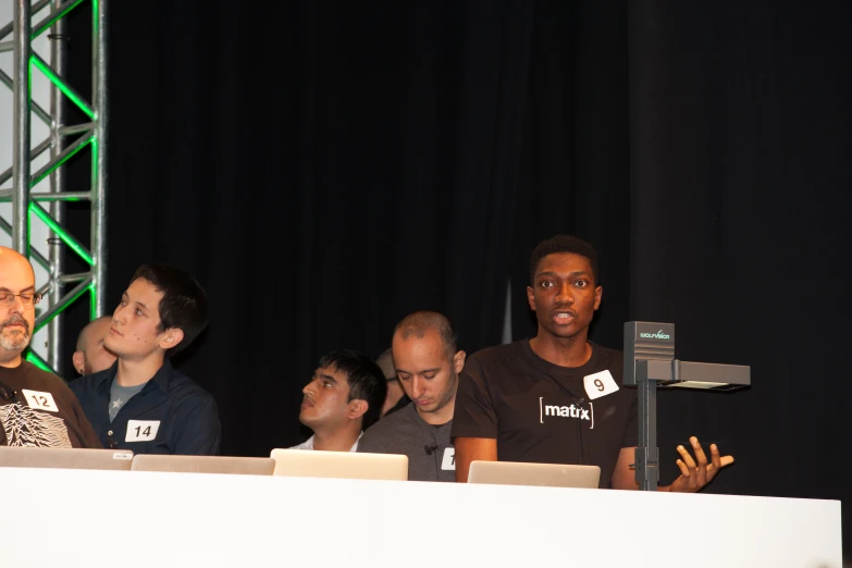 several people sit at tables with laptops while an audience watches