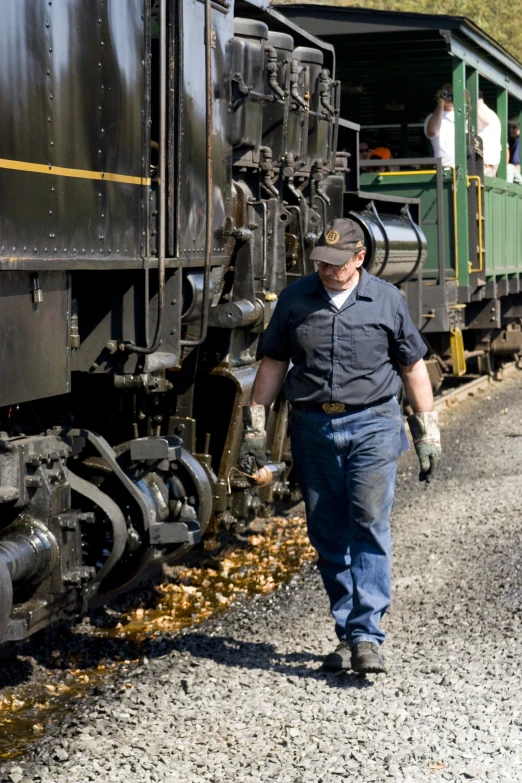 a man in a black shirt walks on a dirt road next to a train with wheels and a yellow stripe down it's side