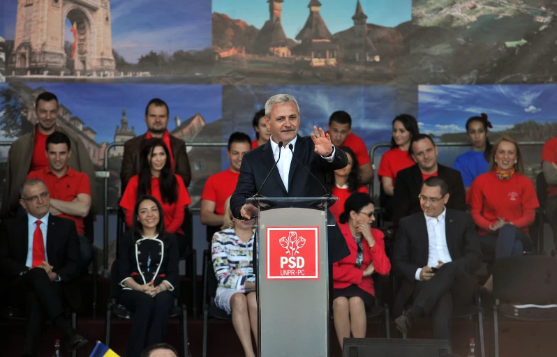 a man speaking at a podium surrounded by people