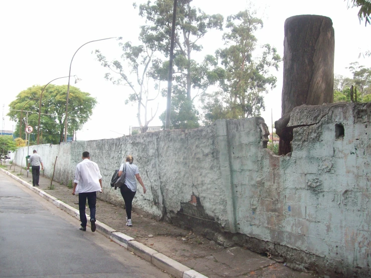 two people are walking down the street, past an old wall