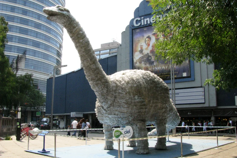 a dinosaur sculpture with people walking by a city building