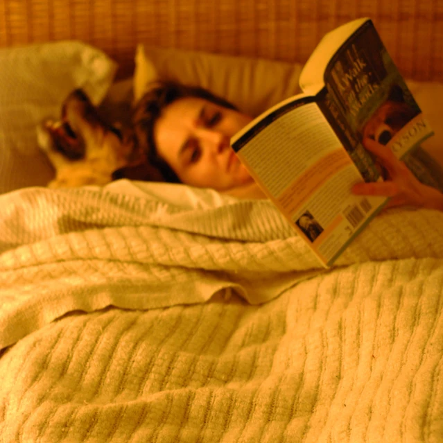 a woman lays in bed with her dog reading a book