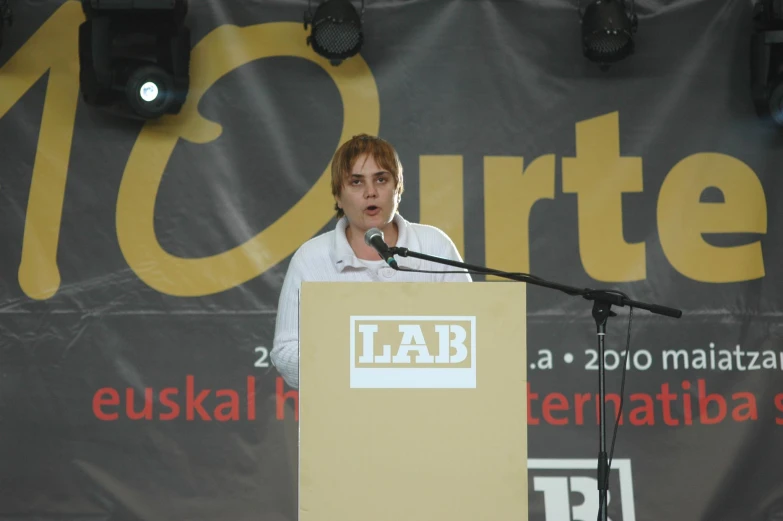 man with red hair speaking at podium in front of wall sign