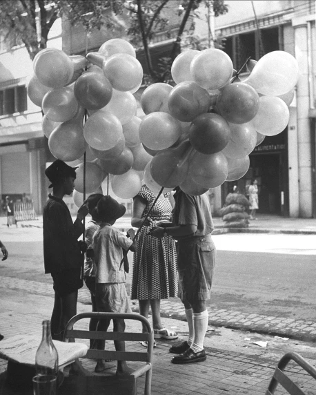 people stand on a city street near a bench and display large balloons