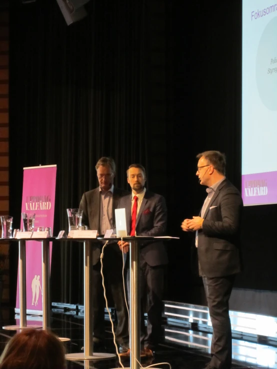 four people standing at a podium, three wearing suits and two talking