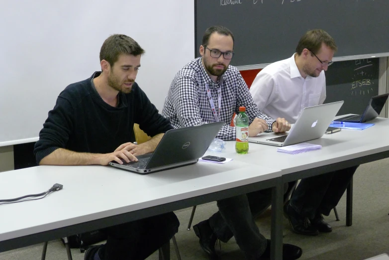 three men sitting at a desk in front of laptops