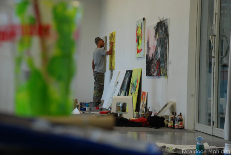 man with paintbrush at the easel in front of wall of paintings