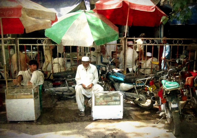 an old man sits under an umbrella beside two bicycles