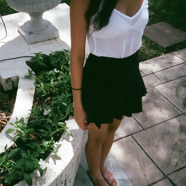 a woman standing near a garden area wearing a skirt and white top