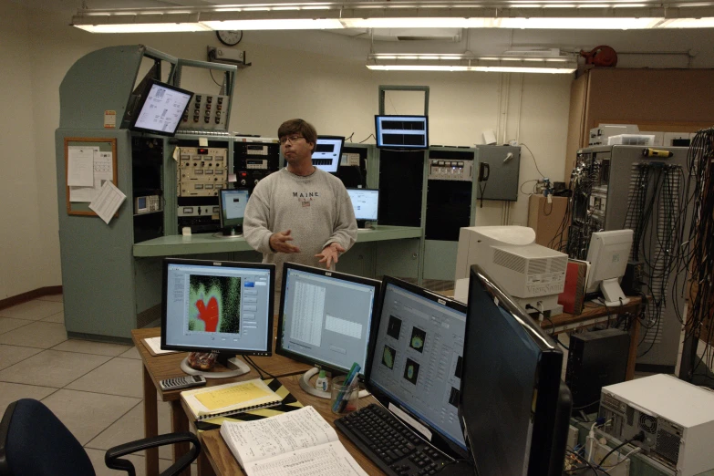 two men stand in front of a desk with multiple computer monitors