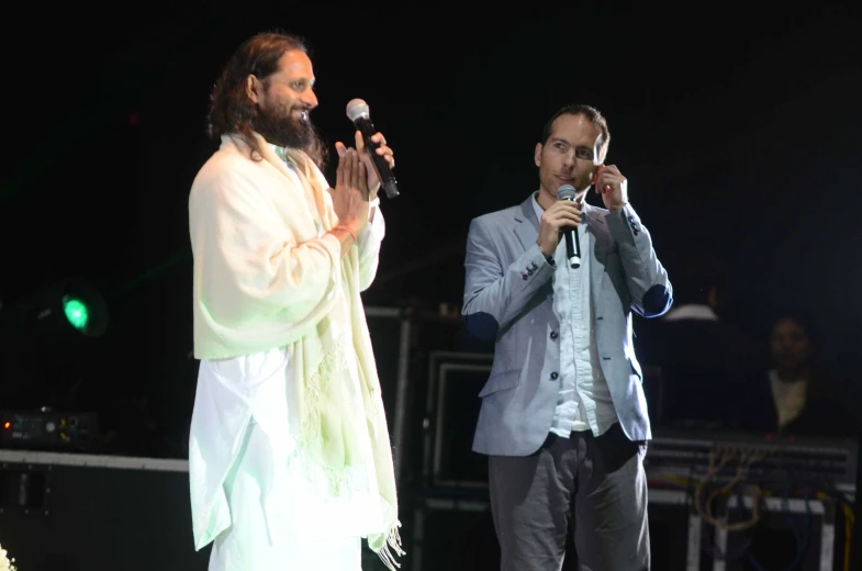 two men stand near each other as one uses a microphone